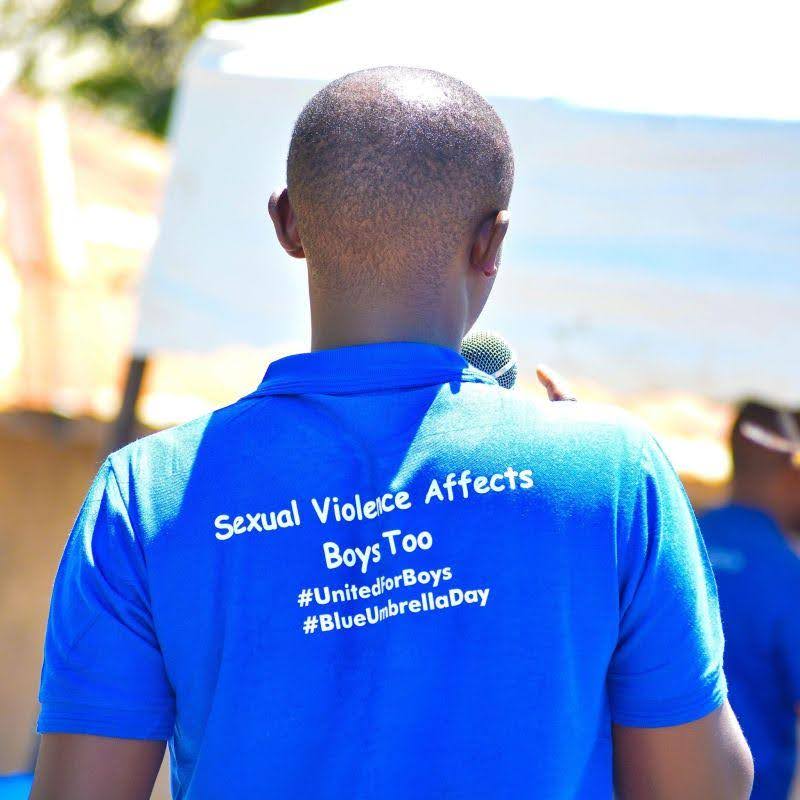 Black man facing backwards using a blue t-shirt. The t-shirt says "Sexual Violence Affects Boys Too" and contains the hashtags #UnitedforBoys and #BlueUmbrellaDay