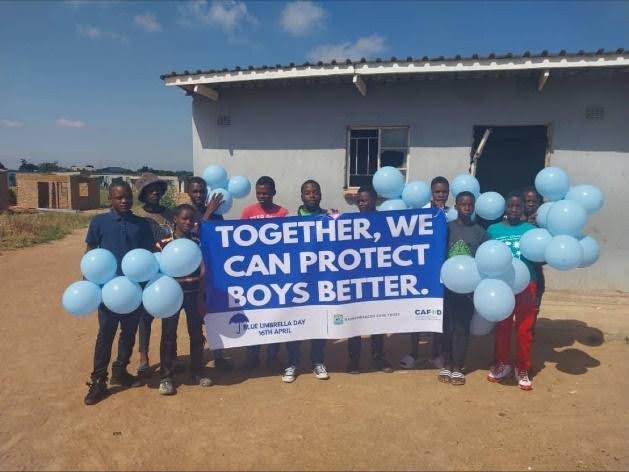 A group of black boys in front of a house, holding blue balloons and a blue and white banner with the saying: "Together, we can protect boys better". The banner has the Blue Umbrella Day logo.